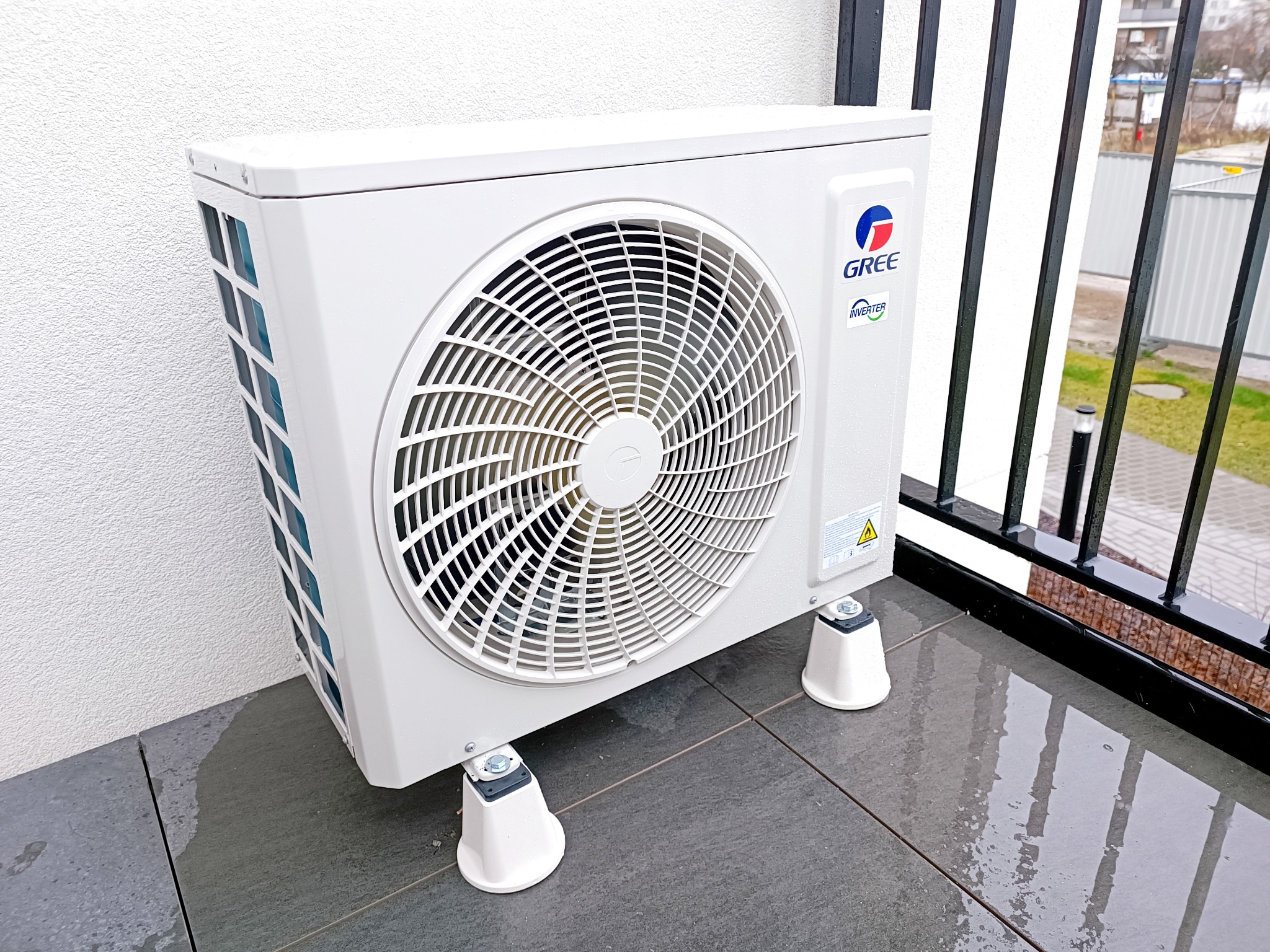 Biała jednostka zewnętrzna klimatyzacji GREE na szarych płytkach balkonu, widoczne mocowania antywibracyjne, fragment balustrady i elewacja budynku w tle.