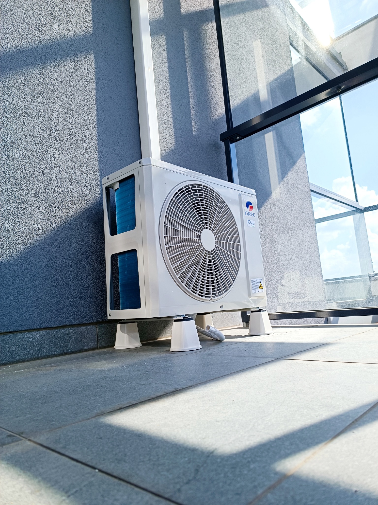 Zewnętrzna jednostka klimatyzacji marki Gree umieszczona na podwyższeniu, widok z dołu, z widocznym fragmentem szarej ściany budynku i przeszklonego balkonu w tle.