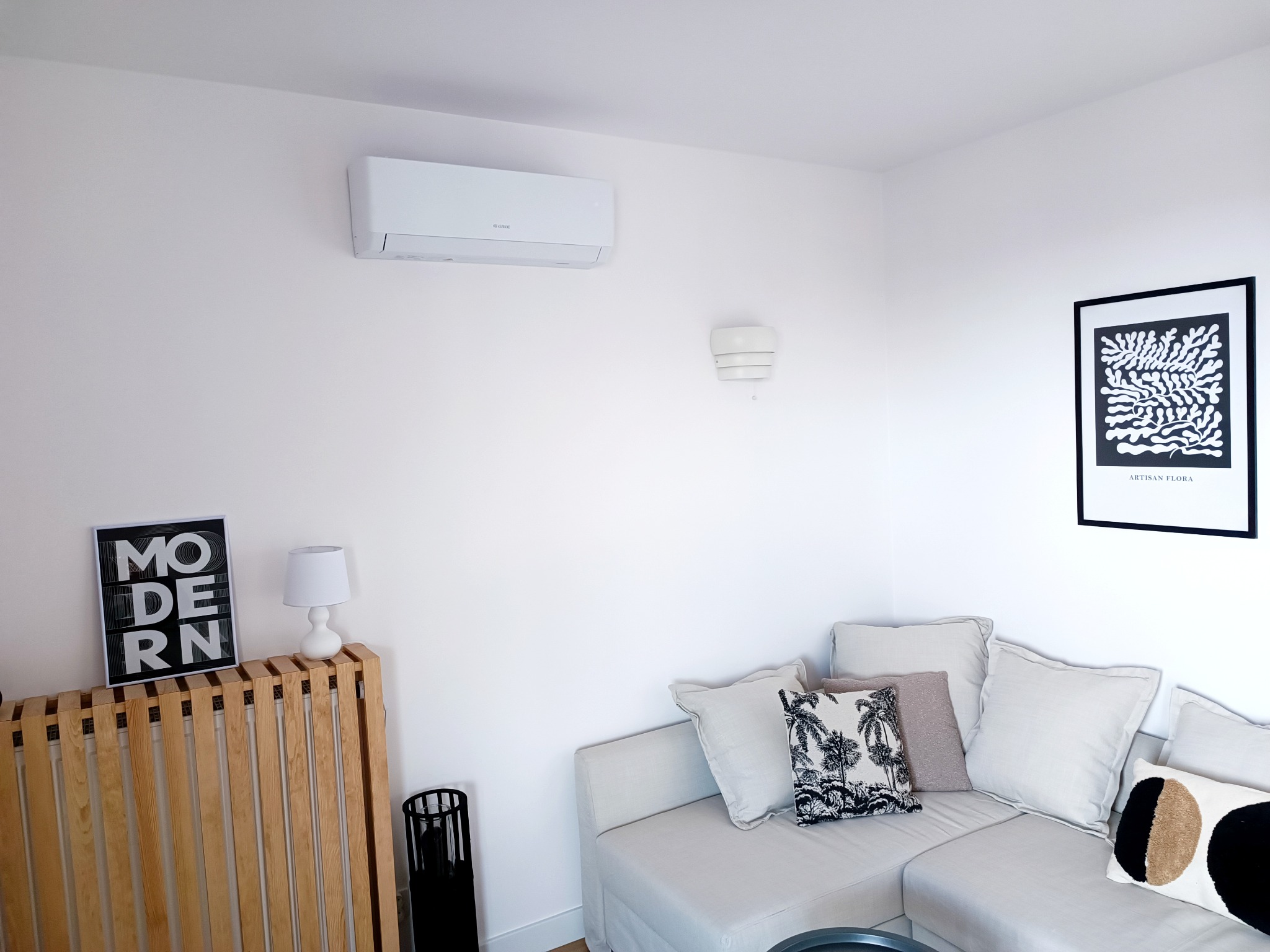 Biały klimatyzator na ścianie w minimalistycznym salonie z jasną sofą, dekoracyjnymi poduszkami, plakatem i drewnianym grzejnikiem z nowoczesnym napisem.