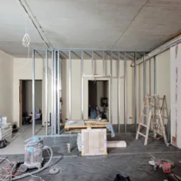 Wnętrze w trakcie prac z metalową konstrukcją ścianki działowej, widoczny surowy betonowy sufit, rozłożona folia ochronna na podłodze, drabina i narzędzia.