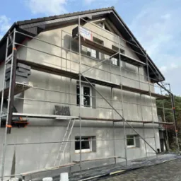 Dom w trakcie ocieplania, widoczne rusztowania, styropian na rusztowaniu, otynkowana fasada, okna w białych ramach, przygotowania do nałożenia elewacji.