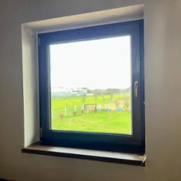 Zamontowane okno z ciemną ramą i drewnianym parapetem w surowym wnętrzu, widok na zielony ogr&oacute;d z elementami dekoracyjnymi.