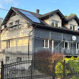 Łukasz Mielan - Dom jednorodzinny w trakcie termomodernizacji z widocznymi rusztowaniami, ociepleniem ścian grafitowym styropianem, panelami fotowoltaicznymi na dachu i ozdobnym kutym ogrodzeniem.