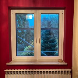 Łukasz Mielan - Dwukomorowe okno z białymi ramami na tle czerwonej ściany i beżowych zasłon, widok na zewnątrz z rusztowaniem w tle.