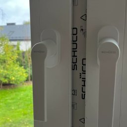 Łukasz Mielan - Zamknięte białe okno z klamkami marki Schüco, widok na ogród i fragment czarnego samochodu za oknem, folia ochronna na ramie okiennej.