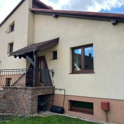 Łukasz Mielan - Dom jednorodzinny z beżową elewacją, brązowymi oknami PCV z widocznymi refleksami nieba, balkonem z czarną balustradą i zadaszeniem, oraz zielonym trawnikiem przed budynkiem.