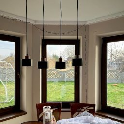 Łukasz Mielan - Wnętrze z trzema oknami drewnianymi w ciemnych ramach, przez które widać zielony trawnik, siatkę do piłki nożnej i ozdobny betonowy płot. Nad stołem wiszą trzy czarne lampy z miedzianymi detalami.