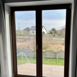 Łukasz Mielan - Widok z okna z brązowymi ramami na podwórko z trawą, betonowym płotem i bramką do piłki nożnej. W tle widoczne domy i pochmurne niebo.