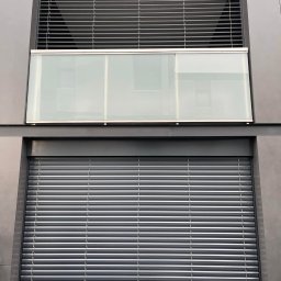 Łukasz Mielan - Elewacja budynku z dwoma oknami: górne z żaluzjami fasadowymi w pozycji otwartej, dolne z roletą zewnętrzną całkowicie zasłoniętą.