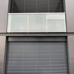 Elewacja budynku z dwoma oknami: górne z żaluzjami fasadowymi w pozycji otwartej, dolne z roletą zewnętrzną całkowicie zasłoniętą.