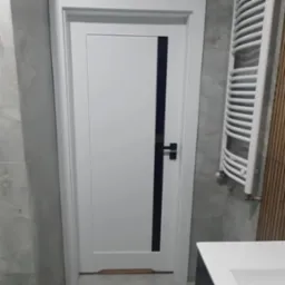 Białe drzwi wewnętrzne z pionowym, czarnym paskiem i czarną klamką na tle szarej ściany i grzejnika drabinkowego w łazience.