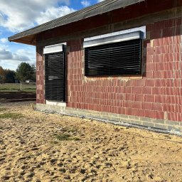 Łukasz Mielan - Dwa okna z zamontowanymi zewnętrznymi przesłonami w kolorze czarnym, widok na surową ceglaną ścianę budynku w trakcie budowy, piaszczyste podłoże.