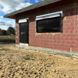 Dwa okna z zamontowanymi zewnętrznymi przesłonami w kolorze czarnym, widok na surową ceglaną ścianę budynku w trakcie budowy, piaszczyste podłoże.