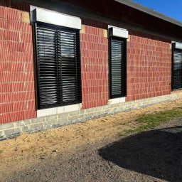 Łukasz Mielan - Elewacja budynku z czerwonej cegły z zamontowanymi czarnymi żaluzjami zewnętrznymi, widoczne także fundamenty i teren wokół budynku.