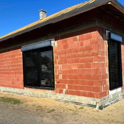 Łukasz Mielan - Surowy budynek z czerwonej cegły z zamontowanymi zewnętrznymi roletami w kolorze czarnym, widoczne fragmenty instalacji drenażowej przy fundamentach.