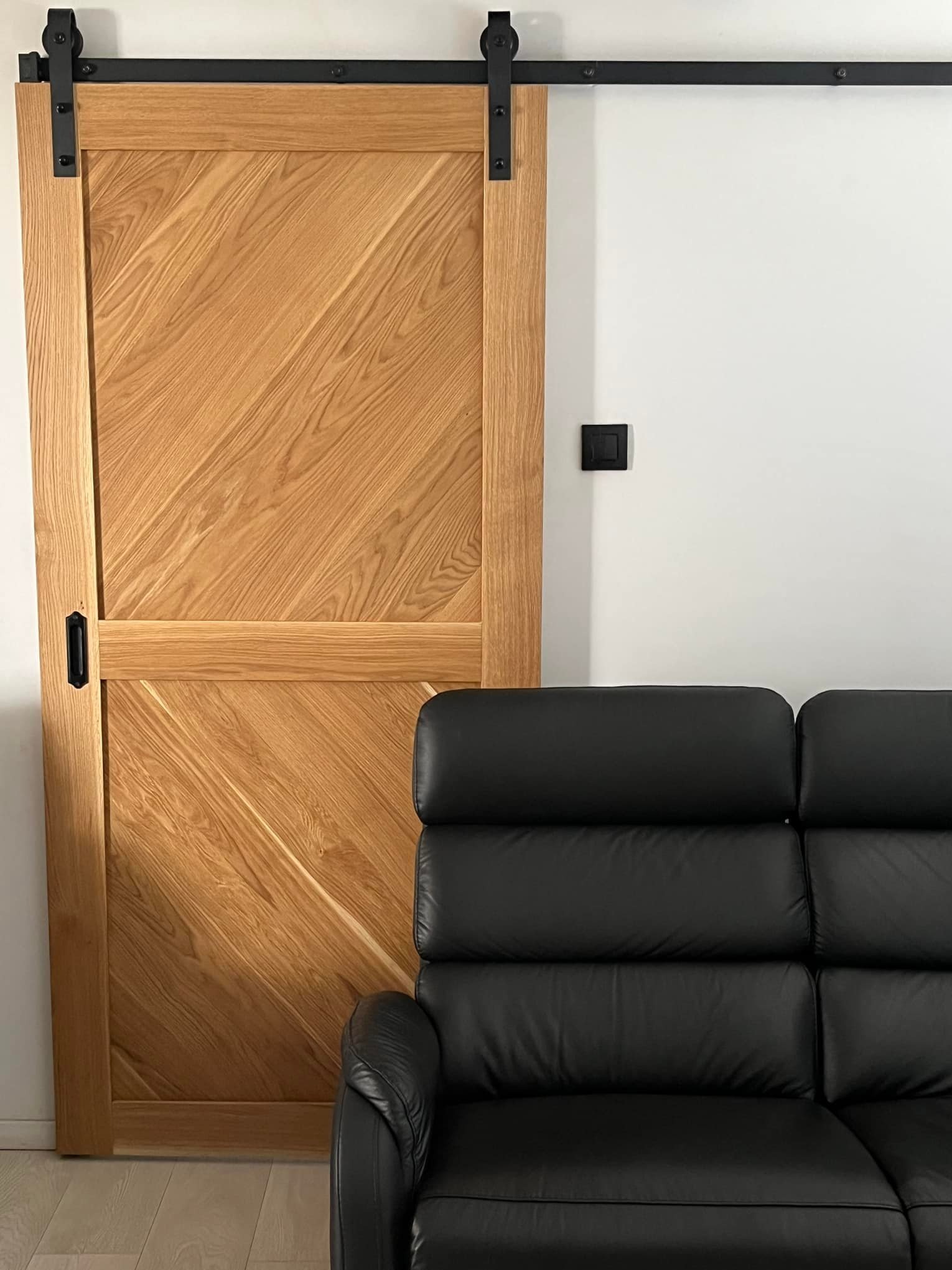 Przesuwne drzwi drewniane w stylu rustykalnym z czarnym systemem szynowym, widoczne obok fragmentu czarnej skórzanej sofy.