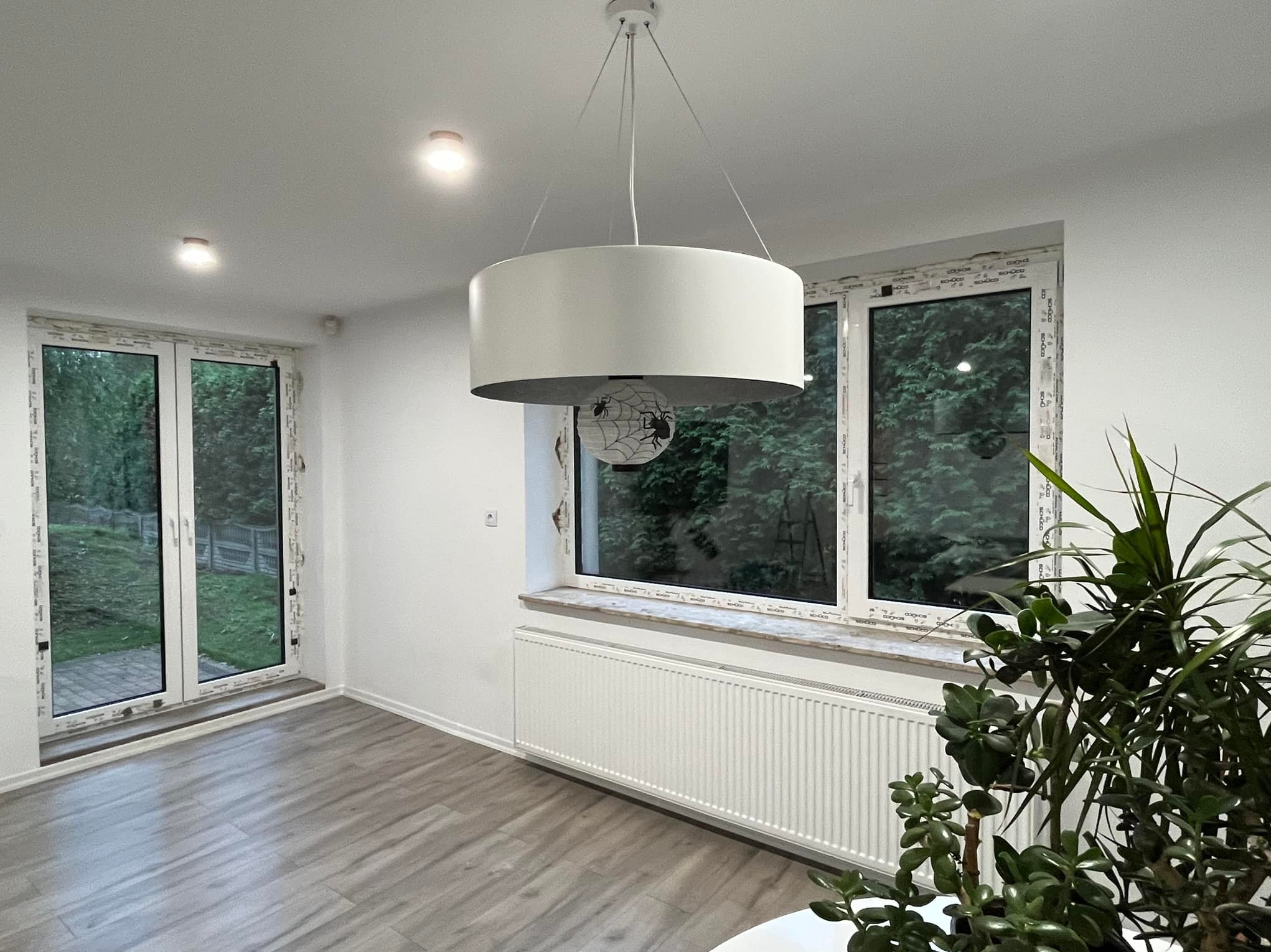 Wnętrze pokoju z dużym oknem, przez które widać zieleń, z białymi ramami okiennymi i białym grzejnikiem pod oknem, na podłodze panele imitujące drewno, w tle drzwi balkonowe z białymi ramami,...