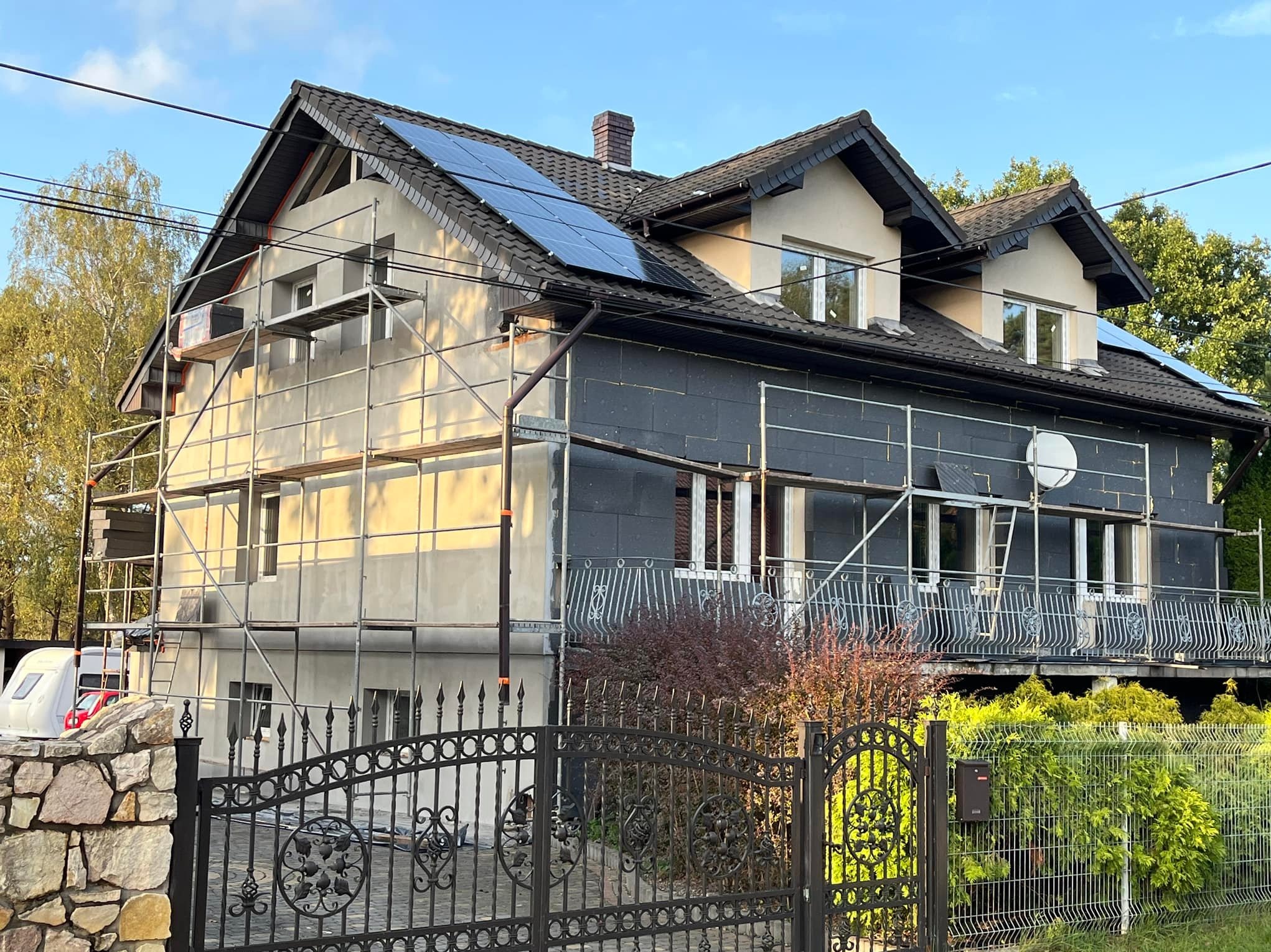 Dom jednorodzinny w trakcie termomodernizacji z widocznymi rusztowaniami, ociepleniem ścian grafitowym styropianem, panelami fotowoltaicznymi na dachu i ozdobnym kutym ogrodzeniem.