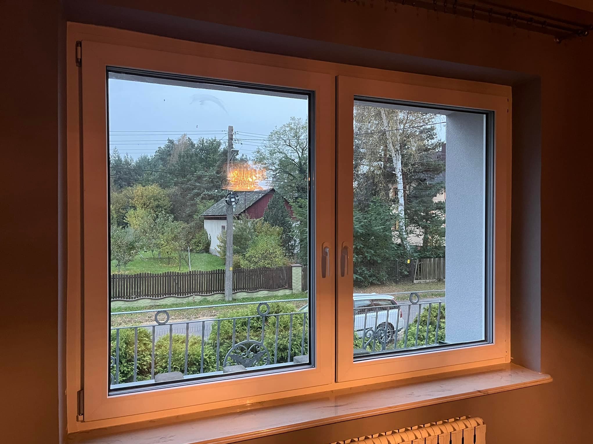 Dwuskrzydłowe okno z białego PCV z widokiem na ulicę z ozdobnym metalowym ogrodzeniem i domy w oddali, uchylone jedno skrzydło.