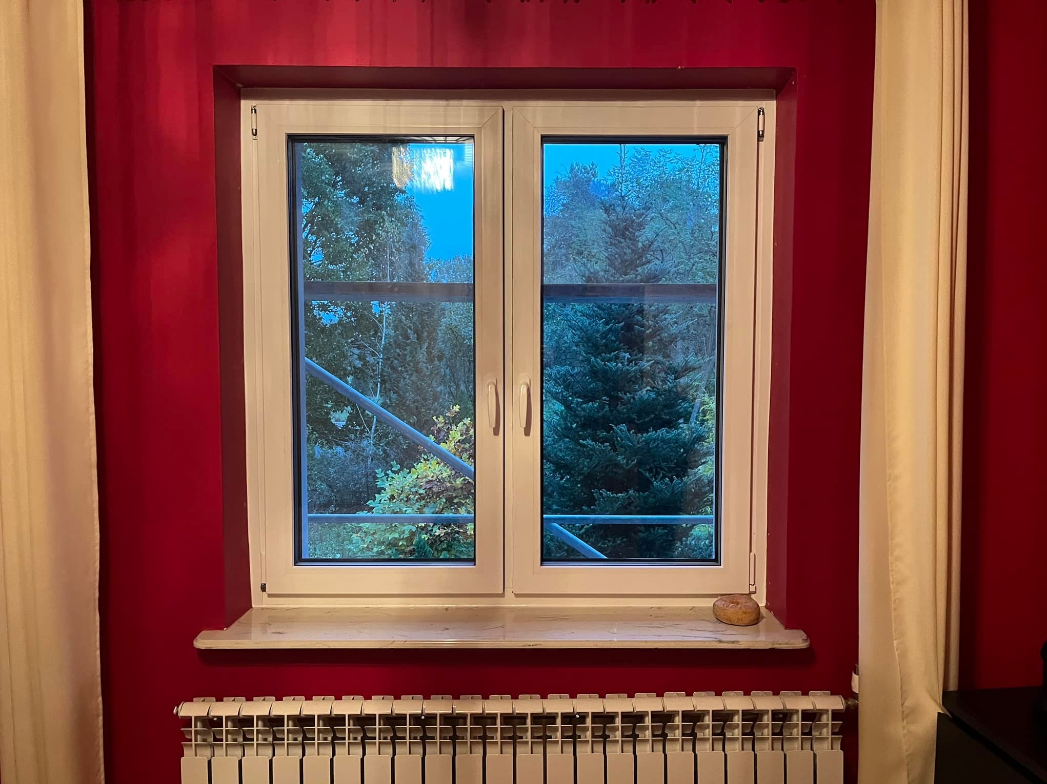 Dwukomorowe okno z białymi ramami na tle czerwonej ściany i beżowych zasłon, widok na zewnątrz z rusztowaniem w tle.