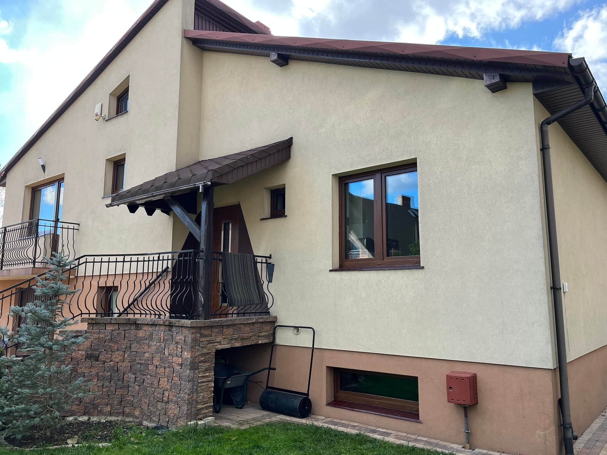 Dom jednorodzinny z beżową elewacją, brązowymi oknami PCV z widocznymi refleksami nieba, balkonem z czarną balustradą i zadaszeniem, oraz zielonym trawnikiem przed budynkiem.
