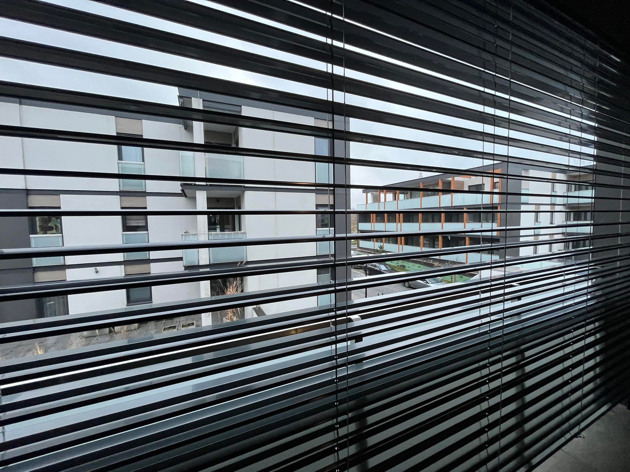 Widok z okna przez częściowo zasłonięte, poziome żaluzje aluminiowe na nowoczesny budynek mieszkalny z balkonami.