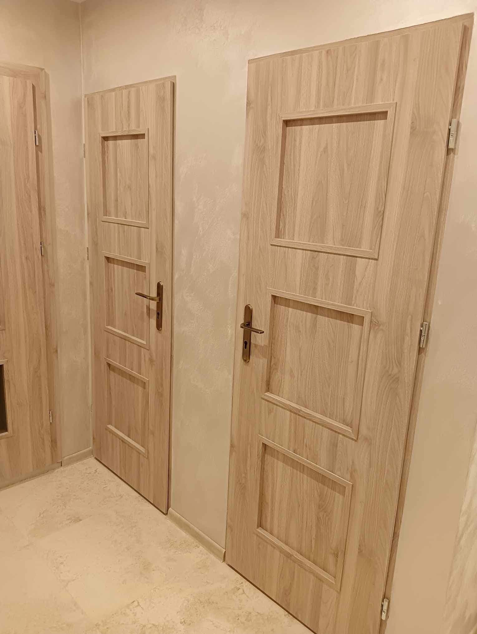 Dwa skrzydła drzwiowe wewnętrzne z jasnego drewna z prostokątnymi płycinami, widoczne klamki i zawiasy, zamontowane w jasnym wnętrzu.