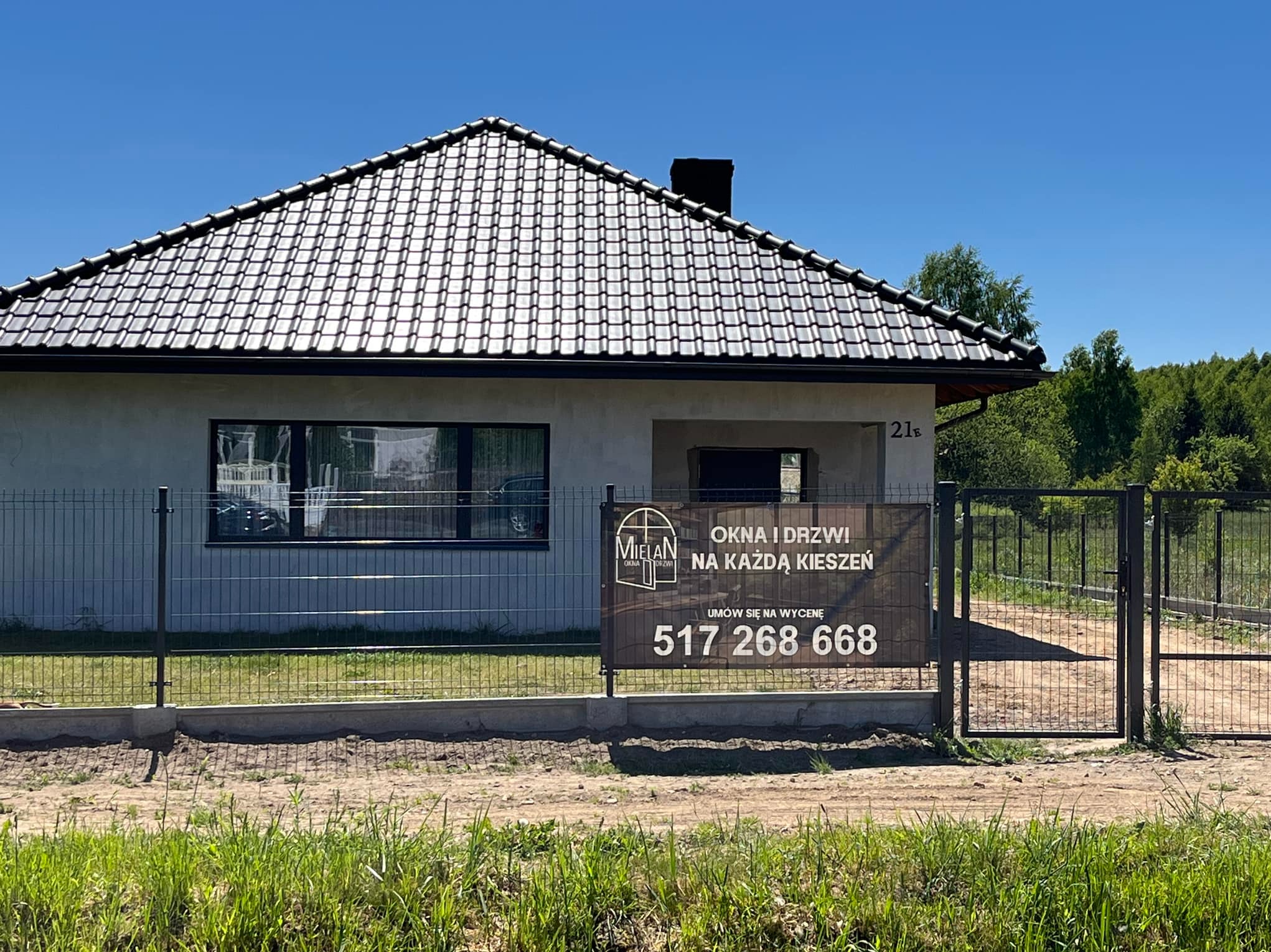 Nowy dom jednorodzinny z szarą elewacją i ciemną dachówką, widoczny przez ogrodzenie panelowe; na ogrodzeniu baner reklamowy firmy oferującej stolarkę okienną i drzwiową z numerem telefonu.