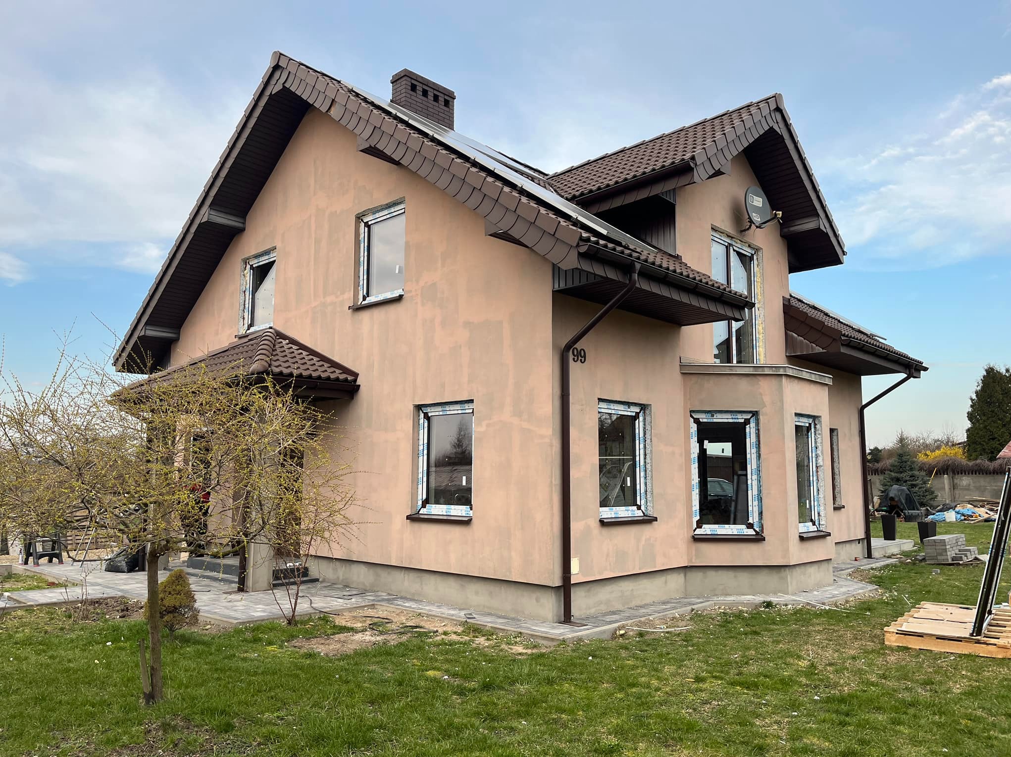 Dom jednorodzinny w trakcie montażu stolarki okiennej, widoczne okna z folią ochronną, elewacja w trakcie wykańczania, w tle drzewa i trawnik.