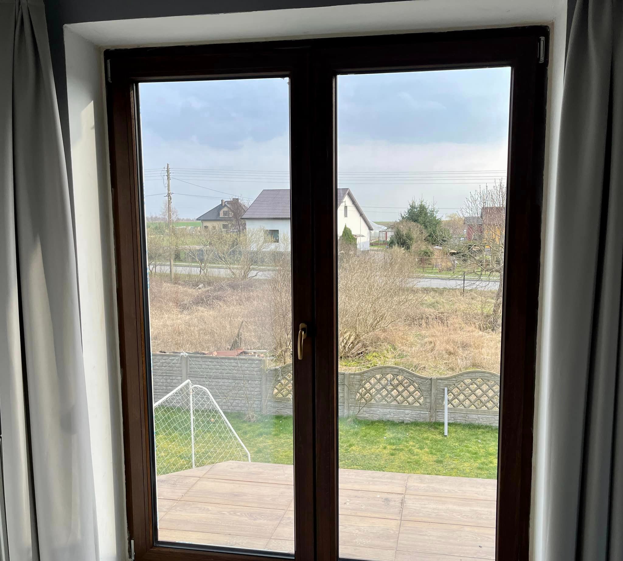 Widok z okna z brązowymi ramami na podwórko z trawą, betonowym płotem i bramką do piłki nożnej. W tle widoczne domy i pochmurne niebo.