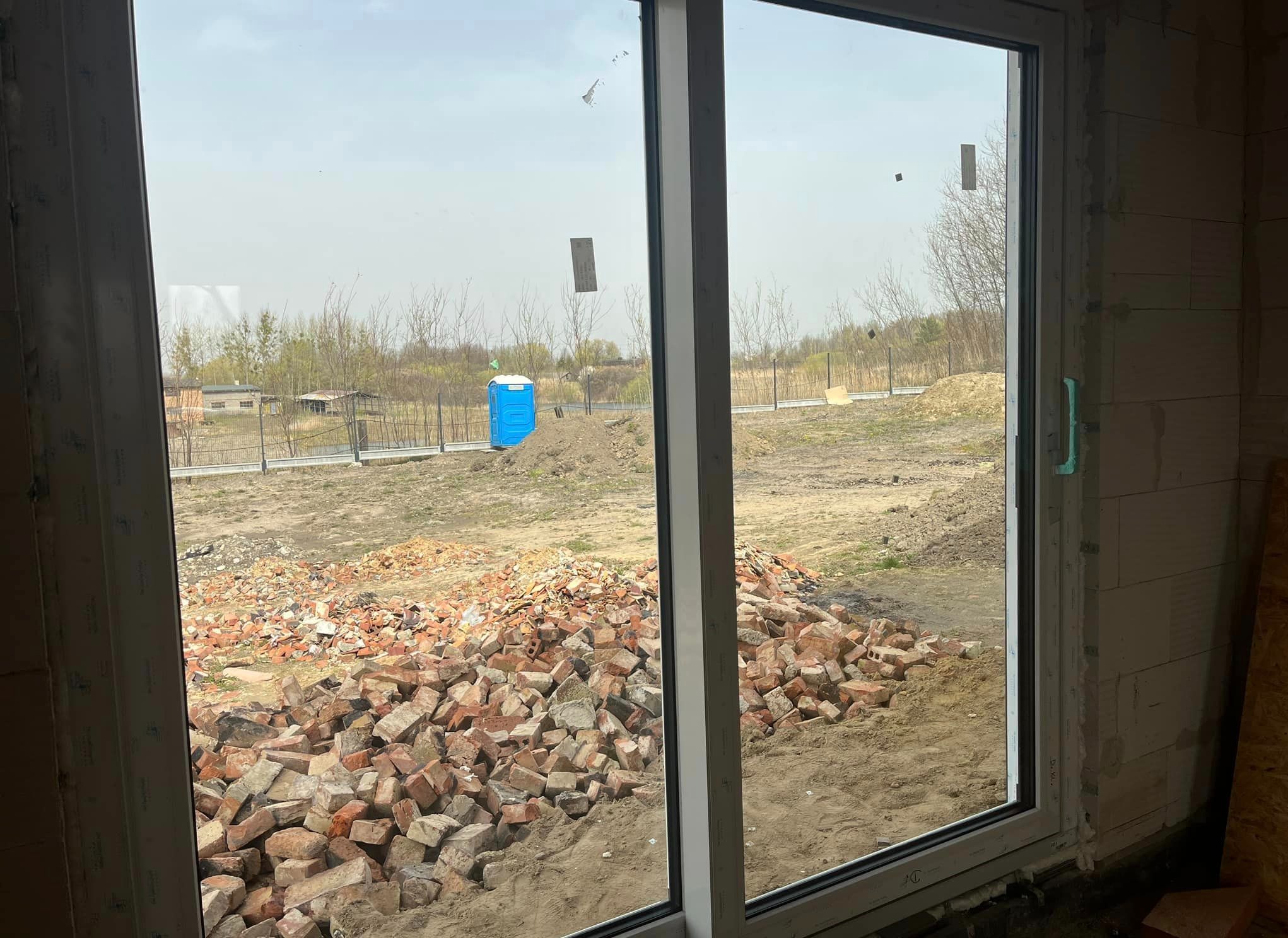 Nowo zamontowane okno z widokiem na plac budowy, gdzie widać stertę cegieł i przenośną toaletę.