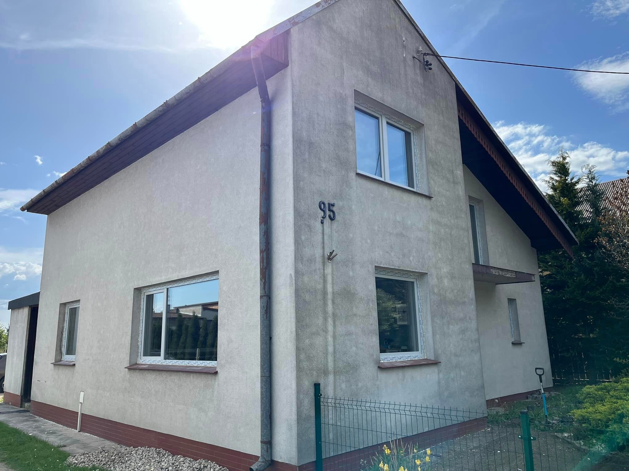 Dwukondygnacyjny dom z numerem 95 na elewacji, z nowymi oknami z białymi ramami, widok z zewnątrz, rynna wzdłuż rogu budynku, ogrodzenie z siatki.