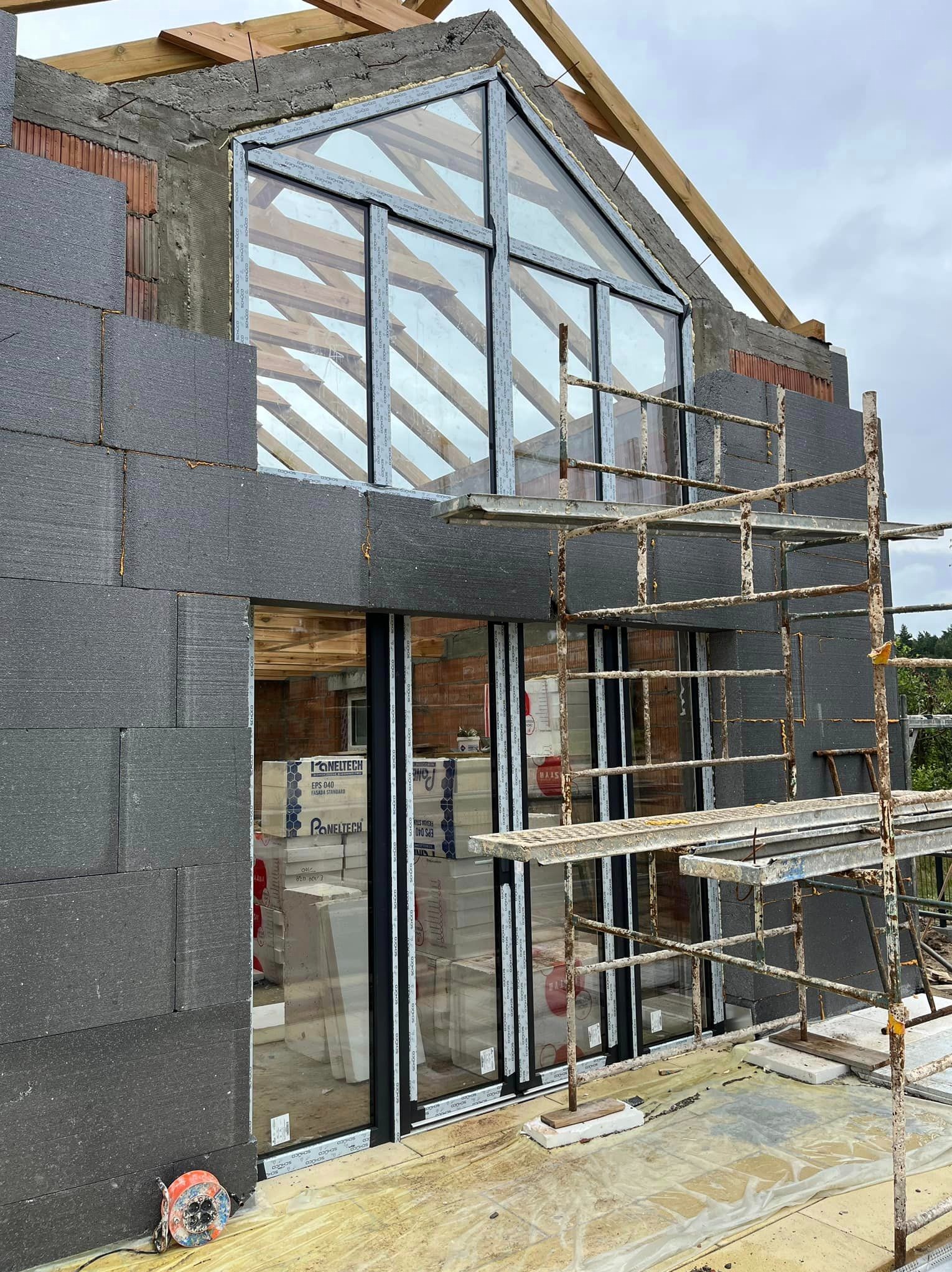 Budowa domu w trakcie montażu stolarki okiennej; widoczne grafitowe bloki izolacyjne, rusztowanie i drewniana konstrukcja dachu.