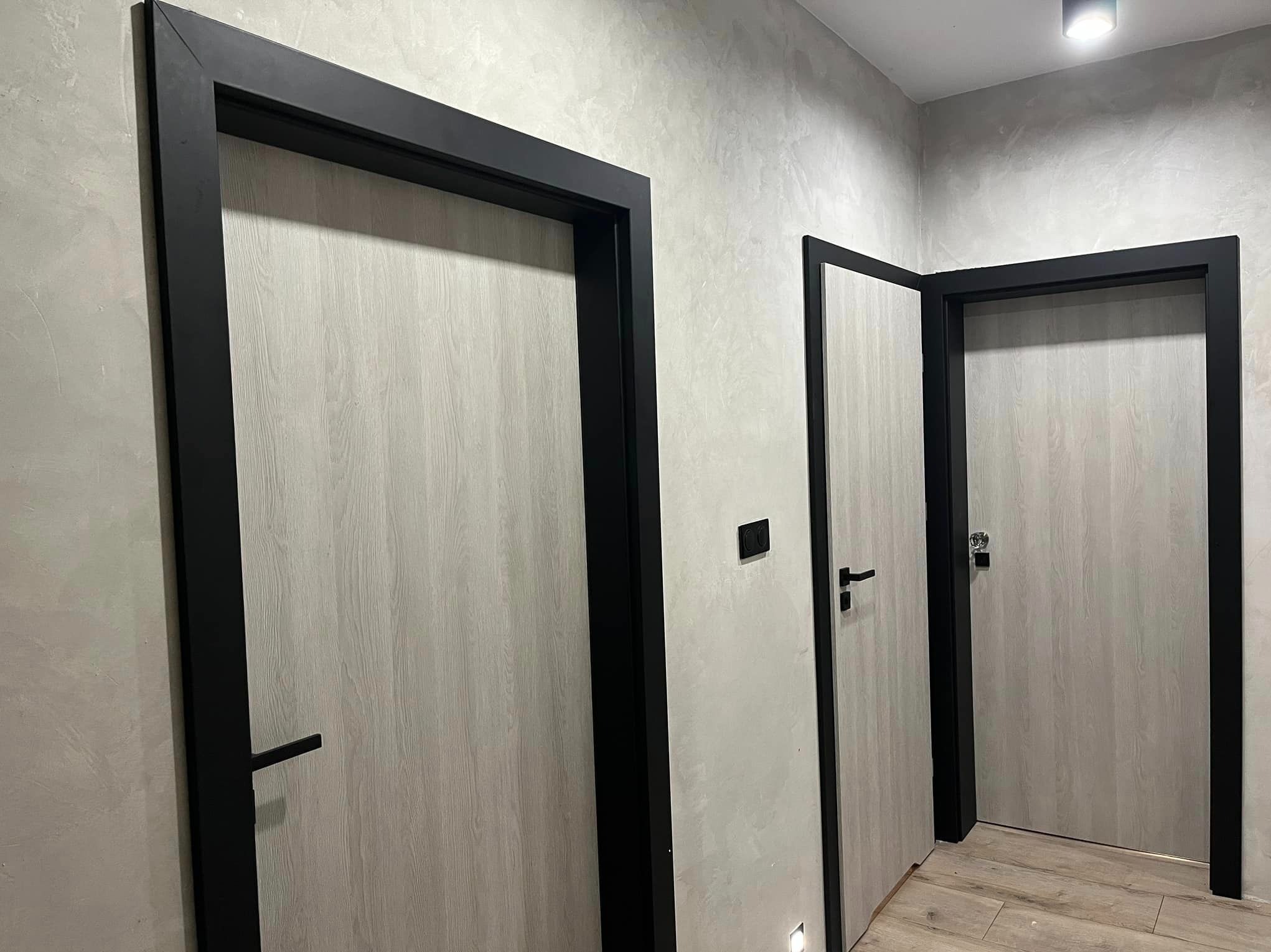 Trzy wewnętrzne drzwi z drewnopodobnym fornirem i czarnymi ościeżnicami w nowoczesnym korytarzu z szarą, teksturowaną ścianą.