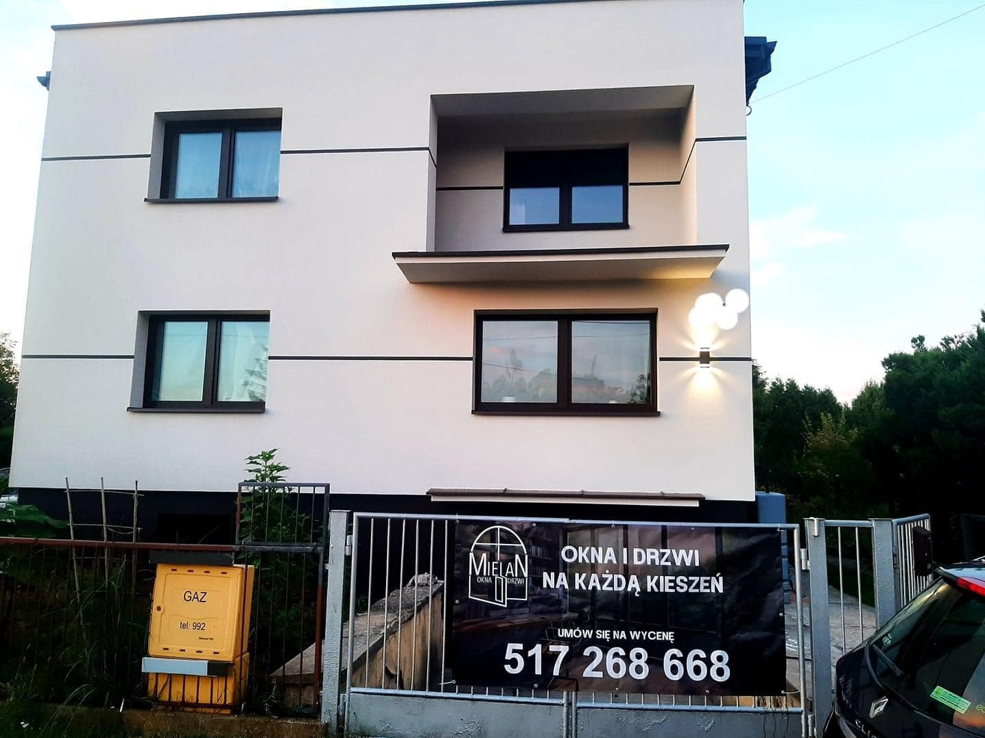 Nowoczesny dom z brązowymi ramami okiennymi i reklamą firmy 'Mielan Okna i Drzwi' na ogrodzeniu, widoczny licznik gazu.