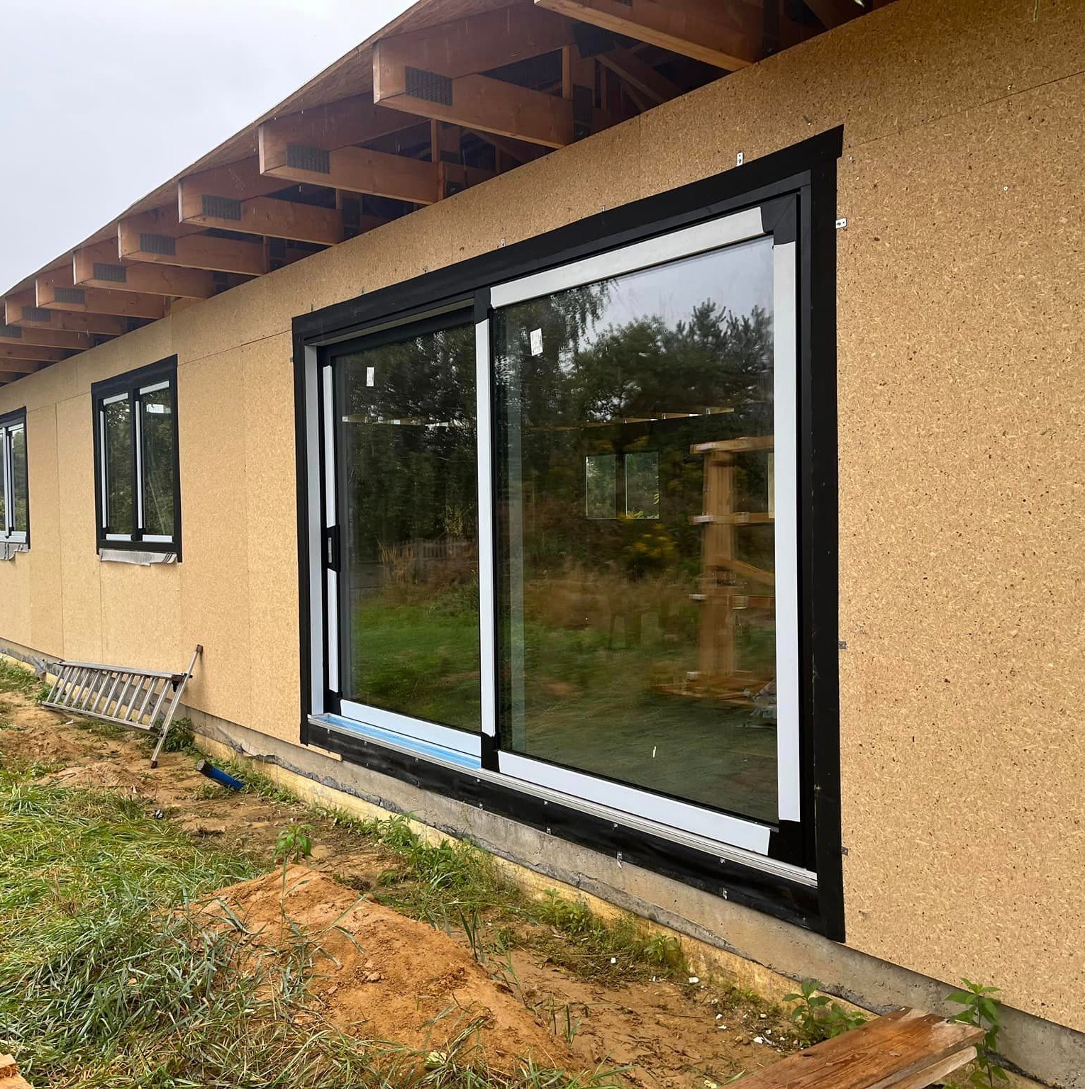 Montaż okna przesuwnego z czarną ramą w surowym stanie budynku, widoczne deski dachu i ocieplenie ścian zewnętrznych.