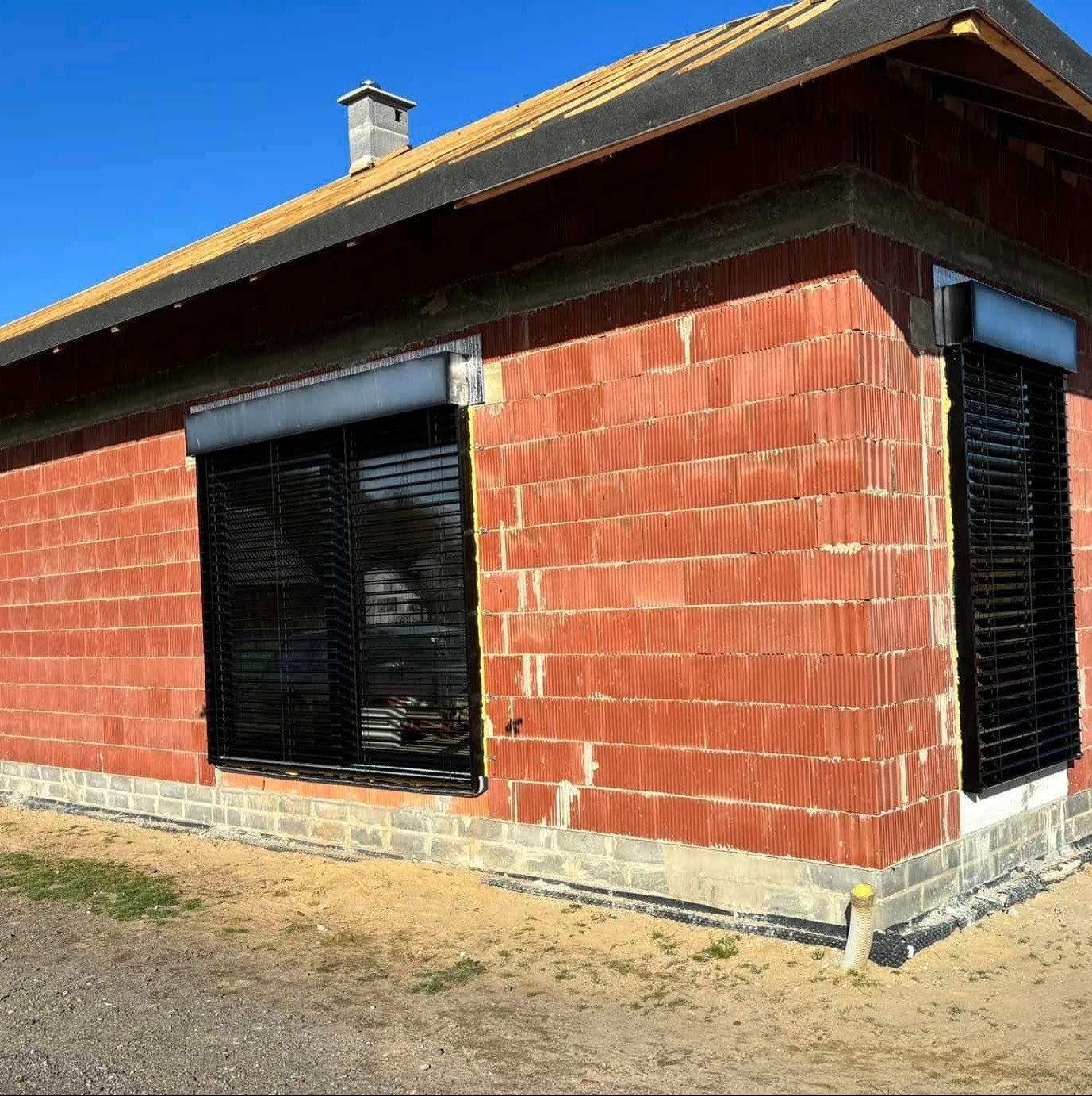 Surowy budynek z czerwonej cegły z zamontowanymi zewnętrznymi roletami w kolorze czarnym, widoczne fragmenty instalacji drenażowej przy fundamentach.