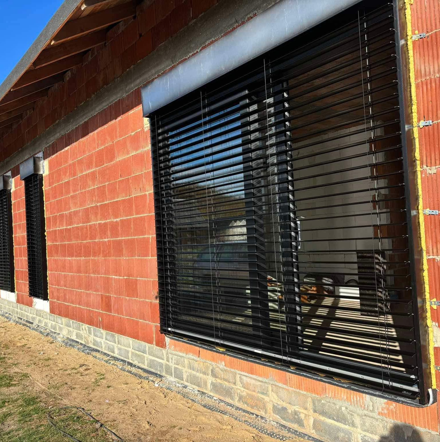 Ceglany budynek w trakcie budowy z zamontowanymi czarnymi żaluzjami fasadowymi, widoczne detale mocowania i surowe wykończenie.