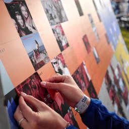 MEMBOX SPÓŁKA Z OGRANICZONĄ ODPOWIEDZIALNOŚCIĄ - Osoba z opaską 'STAFF' na nadgarstku umieszcza zdjęcia z kodami QR na dużej, pomarańczowej tablicy z podziałem na sekcje.