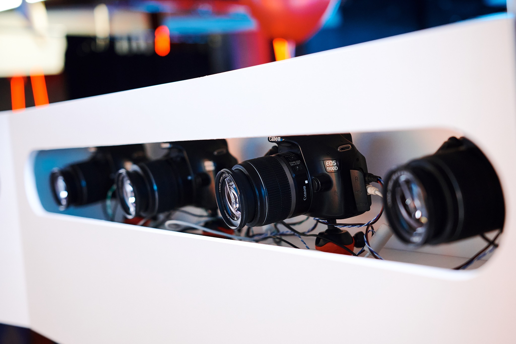 Rząd aparatów Canon EOS 1100D ustawionych w białej obudowie, prawdopodobnie element fotobudki, widoczne kable i statywy.