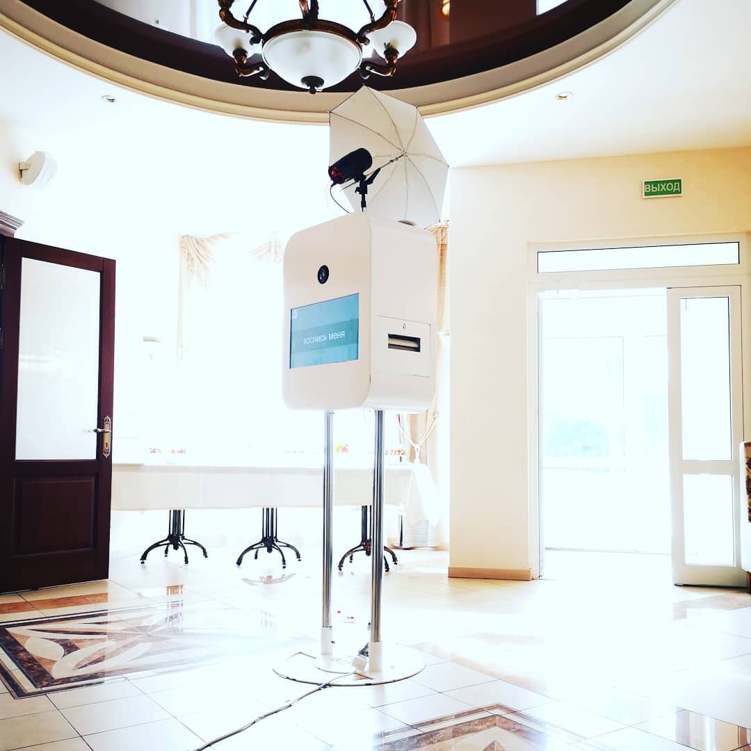 Biała fotobudka z parasolem oświetleniowym w jasnym wnętrzu sali balowej z marmurową podłogą, widoczne stoły nakryte białymi obrusami i otwarte drzwi.