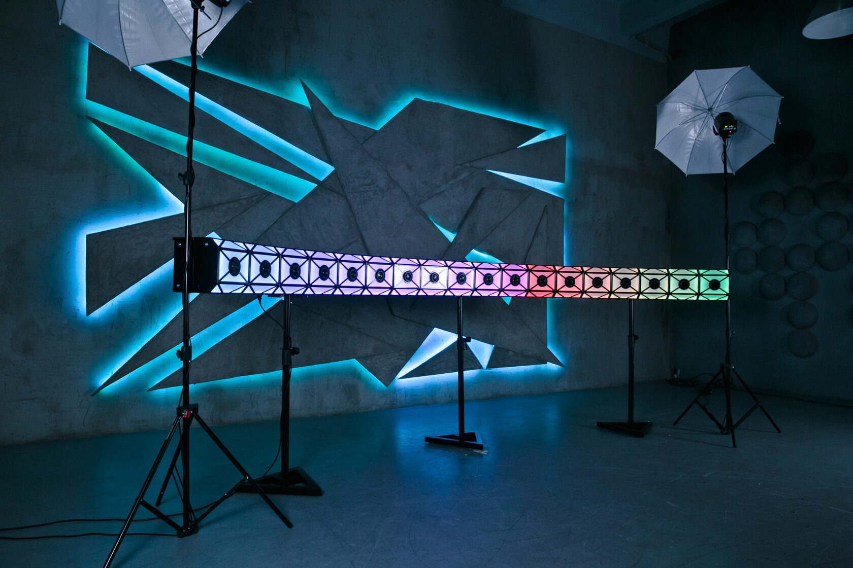 Podświetlana ściana dekoracyjna z geometrycznym wzorem i kolorową listwą LED na statywach, oświetlona parasolkami fotograficznymi.