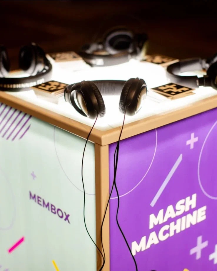 Słuchawki na podświetlanym stole z napisem 'Membox' i 'Mash Machine' na fioletowym tle, otoczone innymi słuchawkami i kartami.