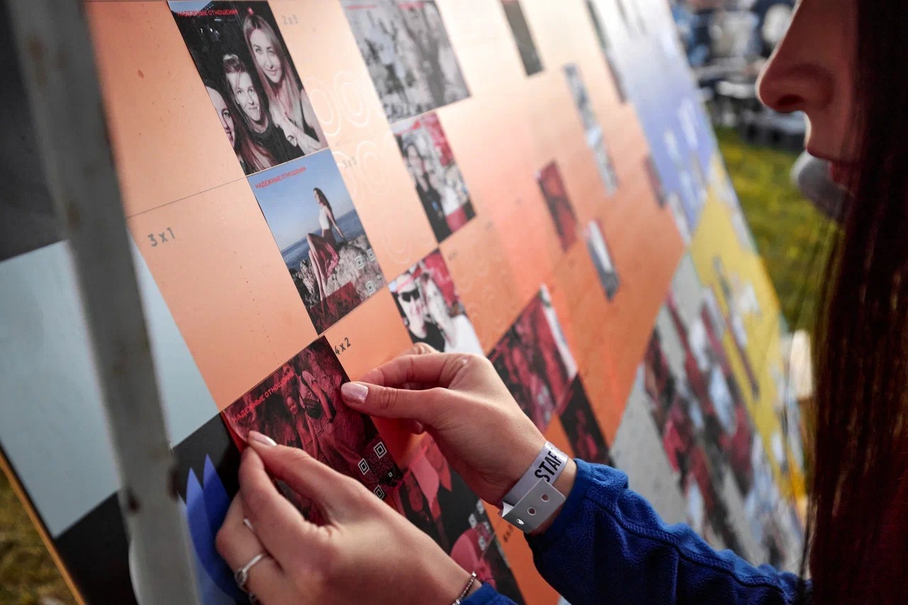 Osoba z opaską 'STAFF' na nadgarstku umieszcza zdjęcia z kodami QR na dużej, pomarańczowej tablicy z podziałem na sekcje.