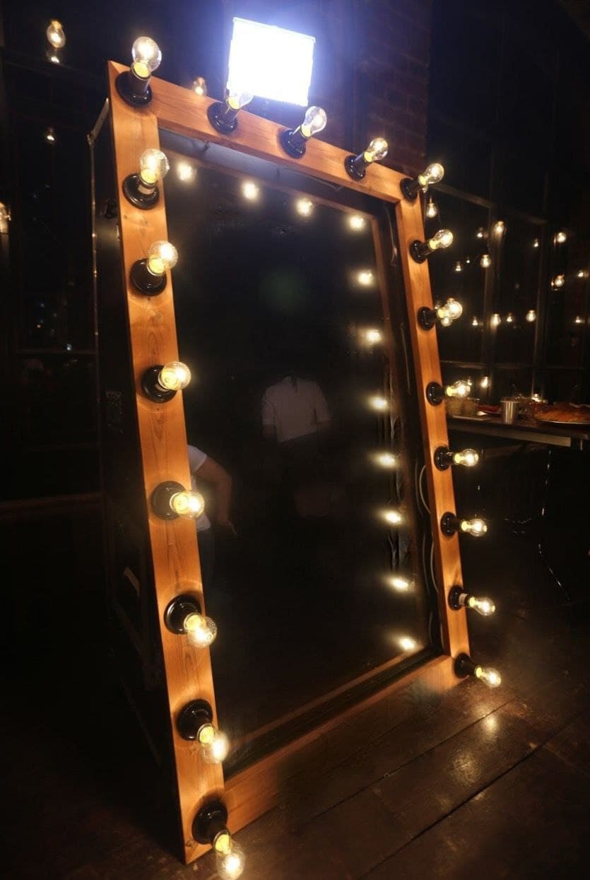 Drewniana fotobudka z lustrem i żarówkami w stylu retro, widoczne odbicia osób i elementy wystroju wnętrza.