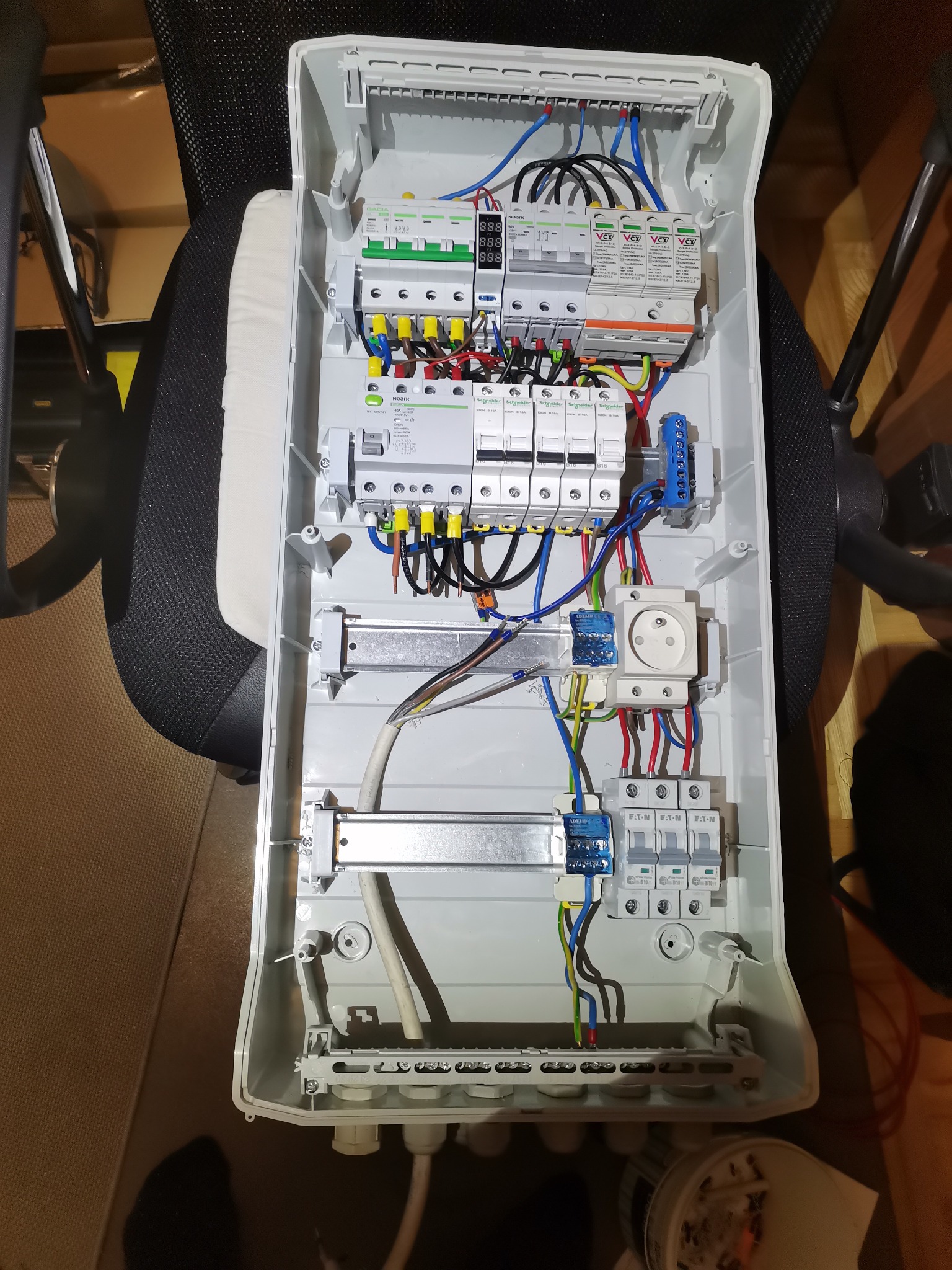 Otwarta, biała skrzynka rozdzielcza z zainstalowanymi bezpiecznikami, wyłącznikami, gniazdem elektrycznym i przewodami o różnych kolorach, widoczna z góry na krześle.