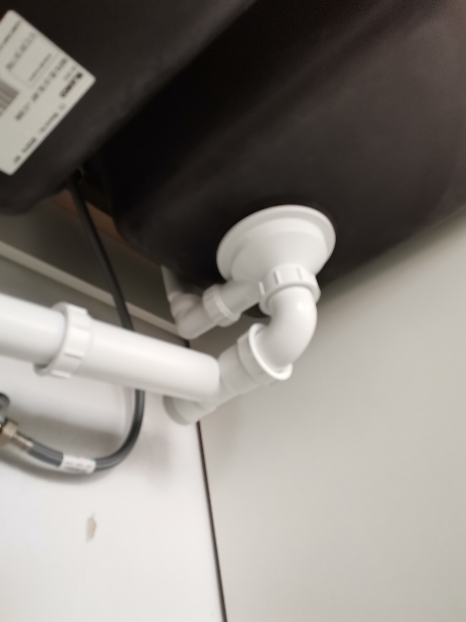 Biały syfon pod zlewozmywakiem, z widocznymi rurami odpływowymi i elastycznym wężem doprowadzającym wodę, zamontowany w szafce z białymi drzwiczkami.