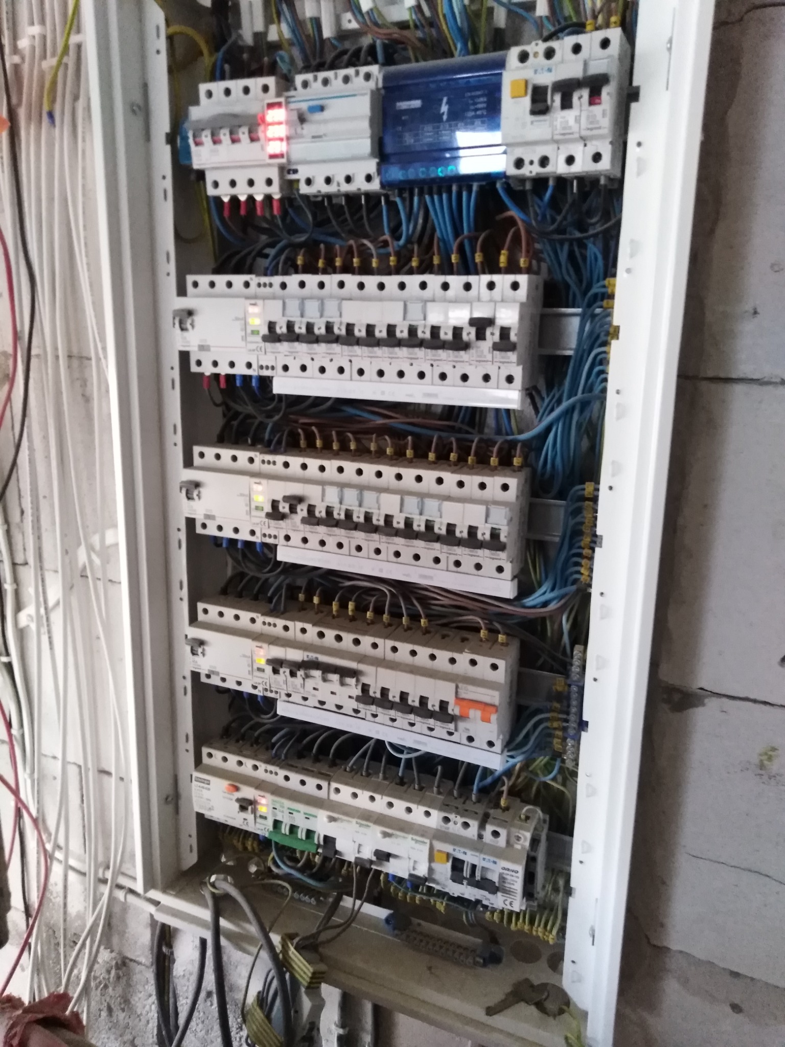 Otwarta skrzynka rozdzielcza z widocznymi bezpiecznikami, przewodami w kolorach niebieskim, brązowym i żółto-zielonym, zamontowana na surowej ścianie z betonu komórkowego.