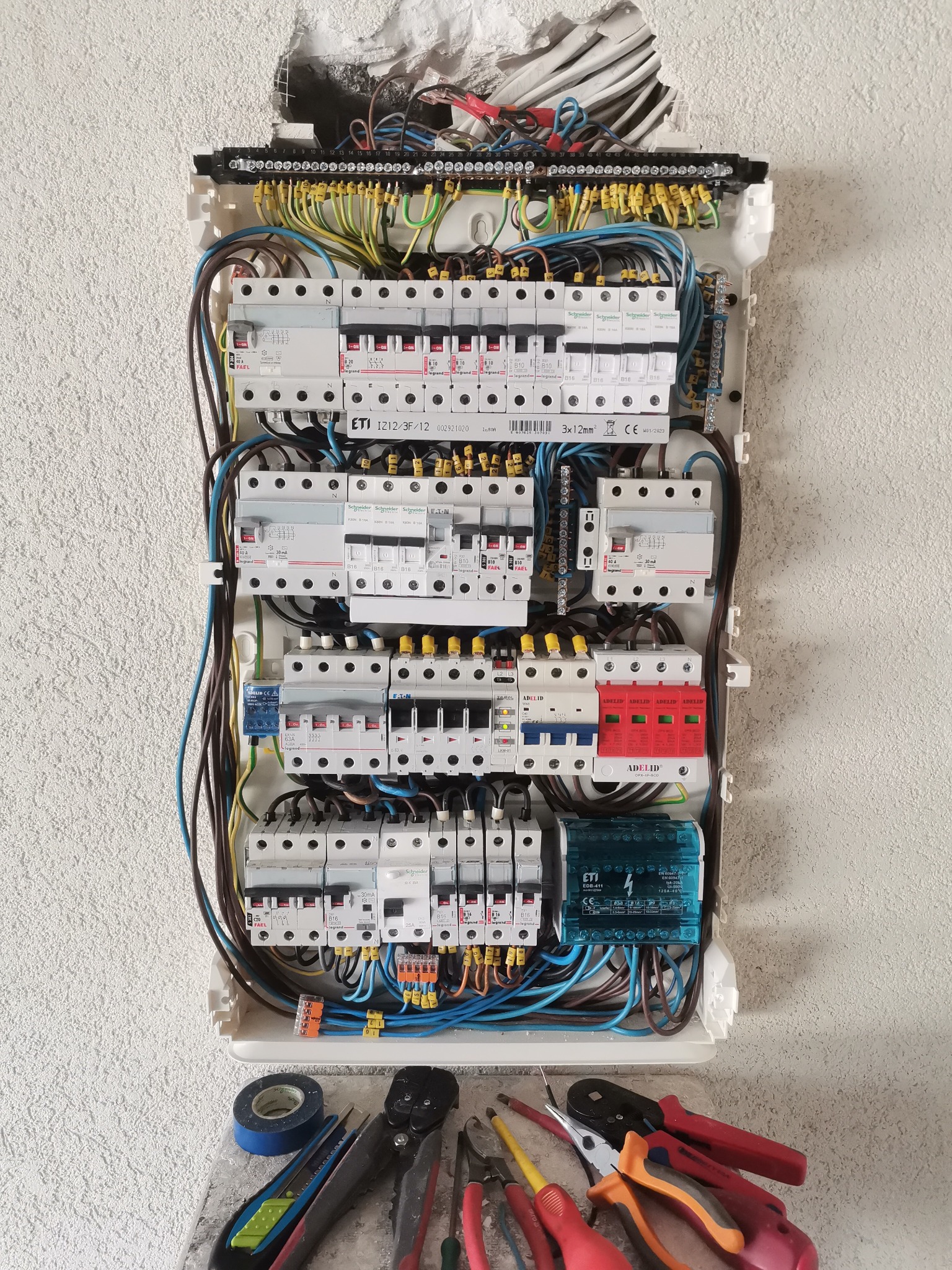 Otwarta skrzynka rozdzielcza z zainstalowanymi bezpiecznikami i przewodami, widoczne narzędzia elektryczne i taśma izolacyjna na tle ściany z widocznym otworem po instalacji.