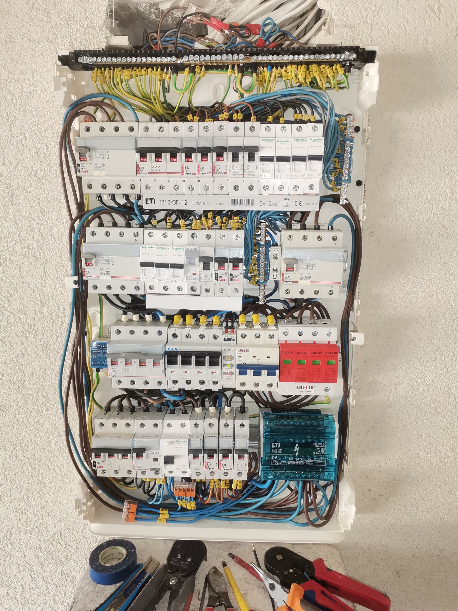 Rozdzielnica elektryczna z zainstalowanymi bezpiecznikami, zabezpieczeniami przepięciowymi i przewodami, uzupełniona o narzędzia na podłodze: taśma izolacyjna, nóż, szczypce, kombinerki.